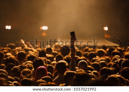 Concerts crowd in music hall.Fan puts hand up with smart phone.Music fans with mobile phones,photography on musical festival crowds.Young man takes picture with phone camera on rock concert.Gig poster