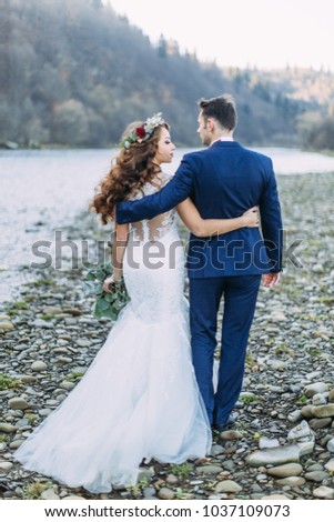Happy bride in a white dress and a wreath of flowers and a groom in a blue suit on the shore of a mountain river