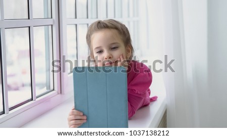 Little girl in a pink dress is sitting by the window with a tablet.