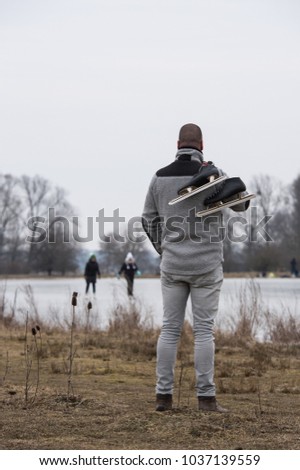 Tall man in wearing grey trousers, a grey coat carrying ice skates looking at other people skating on the ice of a frozen lake (portrait orientation, low depth of field)