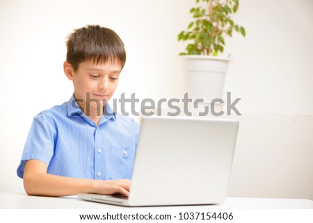 happy boy in a blue shirt uses a laptop sitting indoors at a table. communication, distance education.