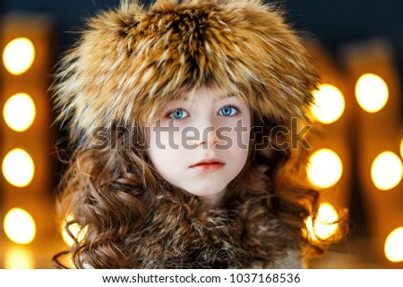 A close up photo of a beautiful little girl with blue eyes and brunette curly hair in studio wearing a fur hat