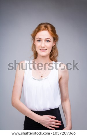 portrait vertical up to the waist young, beautiful business woman, student with lred, curly hair and freckles on gray background. SHe dressed in a white blouse with short sleeves about open shoulders.