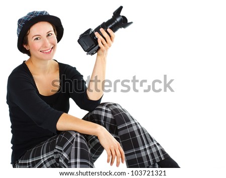 An attractive smiling young woman wearing a hat, black top, black and white checked trousers, sitting and holding up a camera in her left hand