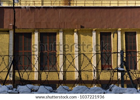 sunlit wall of an industrial building behind a snow-covered fence with barbed wire Royalty-Free Stock Photo #1037221936