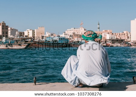 Pakistani or Afghani man in kurta shirt and head wrap sitting and looking out over Dubai Creek with Deira City, abras and boats moored in the background, Dubai, United Arab Emirates