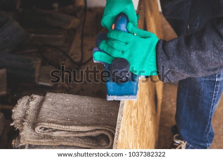 Hands in green working gloves handle the edge of a wooden plate with an electric plane
