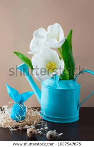 Blue watering can with white tulips and Easter egg in form of rabbit on brown background. Holiday decorating.