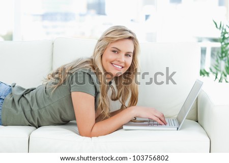 A woman smiling as she uses her laptop, while lying across the couch, and looking forward. Working from home in quarantine lockdown. Social distancing Self Isolation