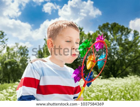 Young boy with toy in hands, blowing with all his might to the windmill on background of spring landscape. Green trees, blue sky with white clouds