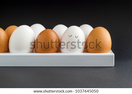 Fresh chicken egg in container on black paper background. Concept for Easter with copy space. White and brown eggs. Painted cute face on a white egg. Drawn cartoon eyes. Frontal view photo.