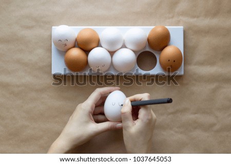 Hands paint cute face on chicken eggs on a brown paper background. Concept for Easter with copy space. Creative layout made of white and brown eggs. Top view photo.