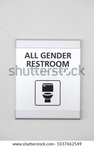 A silver All Gender Restrooms sign with a toilet icon, on a white wall outside of a public bathroom