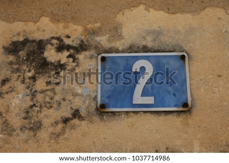 Outdoor exterior view of number two written in white on a blue metallic rectangle. The numeric element is fixed on an old beige and brown wall. Symbol indicating the address in a french city street.