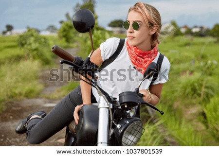 Fashionable young female biker makes stop on long way, stands up from motorbike, wears shades and casual comfortable clothes, has confident look away, rides in countryside. Lifestyle concept