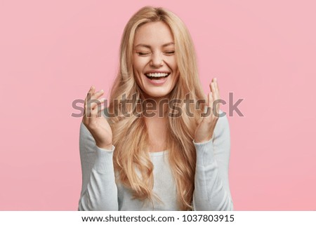 Isolated shot of joyful blonde young cute woman laughs joyfully as hears funny anecdote from friend, has long light hair, poses against pink studio wall. Happiness and positive emotions concept Royalty-Free Stock Photo #1037803915