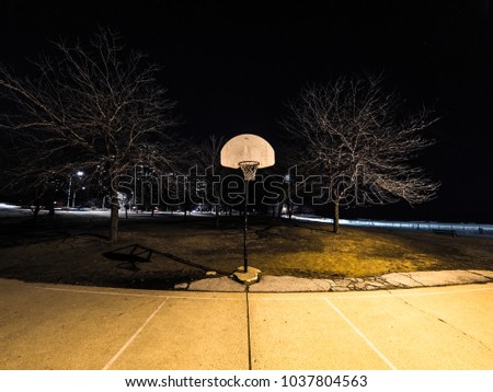 Night photograph of an outdoor weathered basketball court and basketball hoops with white backboards orange rims and nets lit with nearby street light in the city of Chicago.