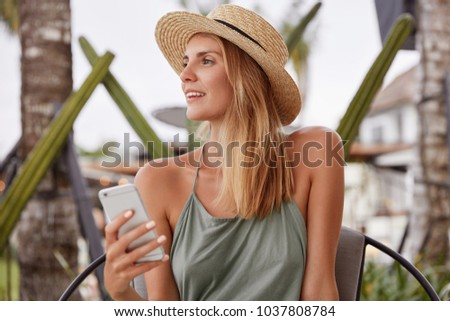Thoughtful adorable woman in casual clothing, keeps modern smart phone, looks aside, recreats outdoor during summer weather, dreams about something pleasant. People, technology, lifestyle concept