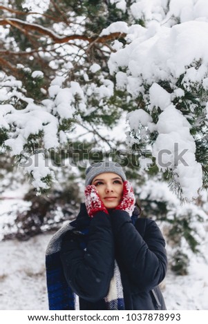 The girl warmes her cheeks in gloves in the cold winter in the pine forest snow-covered