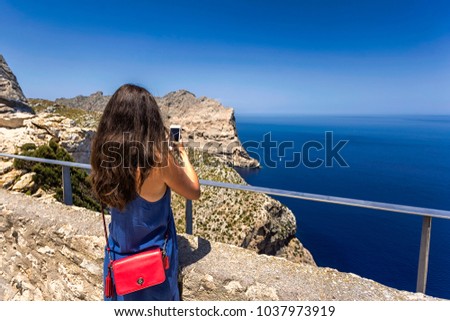 Back view of young woman in a blue dress photographing a view of sea and mountain with her phone