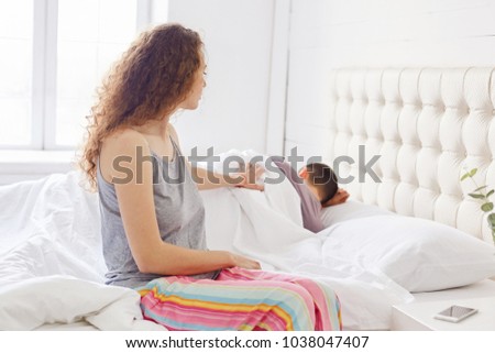 Woman in pyjamas try to wake her husband early in morning, sits on edge of bed, touches him gently with hand, says: Wake up, please! Pretty curly female awakens boyfriend on work. It`s time to get up