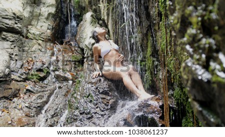 Beautiful young woman in white bikini bathing relaxing under waterfall