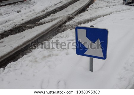 View of a blue and white traffic sign indicating the right hand direction. Dark traces cars on the snowy asphalt road. Abstract urban picture taken in France during the winter. Square element.  
