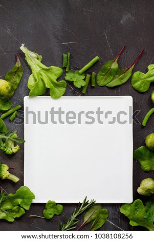 White square plate, various lettuce leaves, rosemary and green vegetables (brussels sprouts, green beans, broccoli) on a dark slate table. Concept of vegetarian food, top view flat lay background.