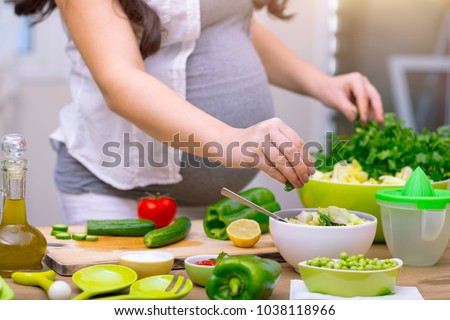 Happy pregnant woman cooking at home, doing fresh green salad, eating many different vegetables during pregnancy, healthy pregnancy concept Royalty-Free Stock Photo #1038118966