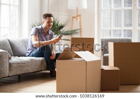 Packed cardboard boxes with young man sitting on sofa in living room calling delivery service at background, belongings in carton containers waiting for moving in out new home or relocation concept Royalty-Free Stock Photo #1038126169