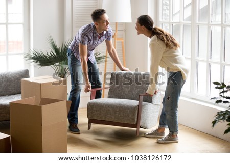 Smiling couple carrying modern chair together placing furniture moving into new home, young family discussing house improvement interior design while furnishing living room, remodeling and renovation Royalty-Free Stock Photo #1038126172
