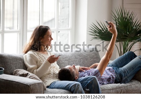 Smiling young millennial couple having fun with smartphones using apps at home making photo or shooting blog on cellphone, happy man taking selfie on mobile phone relaxing with girlfriend on couch