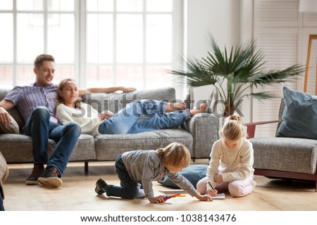 Children sister and brother playing drawing together on floor while young parents relaxing at home on sofa, little boy girl having fun, friendship between siblings, family leisure time in living room Royalty-Free Stock Photo #1038143476