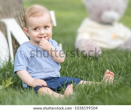 A smiling beautiful small baby boy holding a finger in his mouth sitting on green grass outdoor at summer park. Emotions, smile, surprised, kid, toddler, sly, shy, toy.