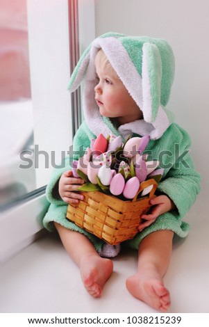 cute kid in an Easter bunny costume with a bouquet of flowers sits on a window sill against the window and smiles. March 8, Easter, spring.