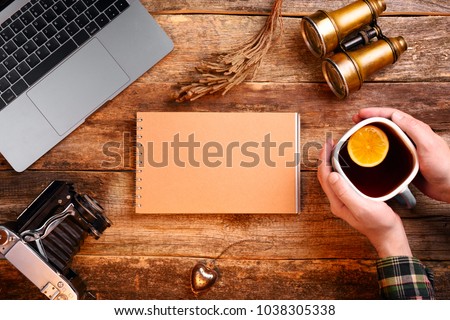 Notepad with blank page of kraft paper in the center of the frame. Old wooden table. Hands holding a cup of tea with a lemon. Binoculars, old camera, notebook, laptop. Mockup. 