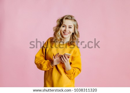 portrait of cheerful  caucasian female with curly blonde hair, poses on a pink background. woman looks for information on the Internet and looks through the websites by means of the smartphone. 