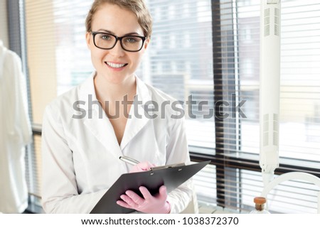 Waist-up portrait of pretty young doctor wearing white coat and rubber gloves distracted from taking notes in order to pose for photography