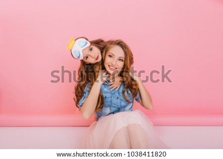 Cute girl in sleep mask lovingly embraces her cheerful mother sitting on the floor on pink background. Adorable young curly mom and long-haired daughter having fun and spending time together