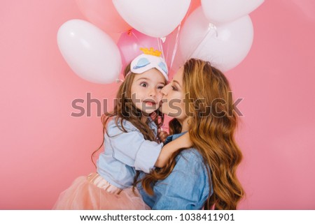 Attractive curly young woman with party balloons congratulates daughter on birthday and kisses her on the cheek. Little girl in sleep mask with surprised face expression embracing mom at event