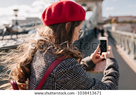 Portrait from back of stylish woman in tweed jacket holding phone and making photo of city landscape. Elegant girl in beret using smartphone while taking picture of town views in travel.