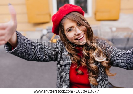 Lovable dark-haired female model in trendy glasses making selfie near yellow building. Close-up portrait of refined cute girl in red hat having fun outdoor.