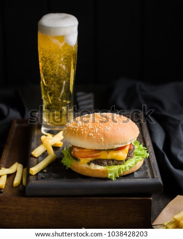 Delicious fresh tasty burger with beef, tomato, cheese and lettuce, french fries and beer served  on a wooden cutting board on dark background.
Street fast food