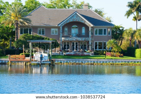 Beautiful two story waterfront house with artistic stone work with palm trees, colorful landscaping and lush green grass in tropical climate with boat on a lift and a FOR SALE sign in the yard.
