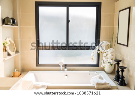 An interior picture of luxury beige bathroom with a white bathtub and white towel with a white artificial flowers in a ceramic pot with a windows and frame.