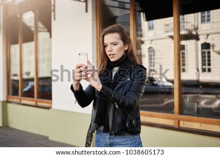Friends will love this photo. Creative modern businesswoman in trendy leather coat holding smartphone while taking picture of scenery or band that plays on street, being entertained and focused