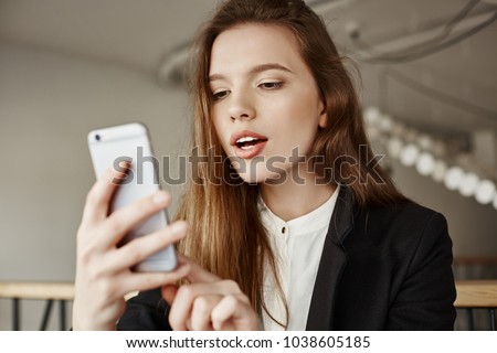 Girl puts filter on her selfie. Indoor shot of stylish modern european student sitting in cafe, updating her profile picture in social network via smartphone, editing it in app. Woman checks lipstick