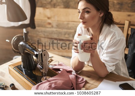 Someday my fashion line become famous. Dreamy female tailor thinking and drinking coffee, sitting near sewing machine and fabric, having break while creating new garment. Creative prefers not to rush