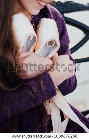 Smiling girl in violet dress with long dark hair is holding pink ballet shoes in hands