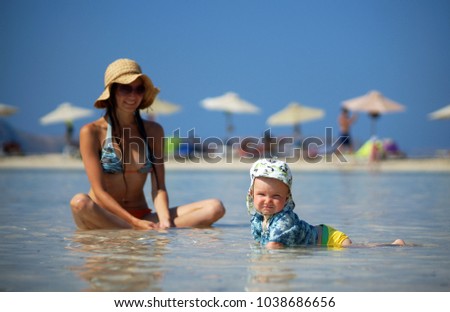 Little smiling child in beach clothes lying in sea shallow water in the foreground. On background young woman in bikini, straw hat and sunglasses sitting in water, sun umprella and beach